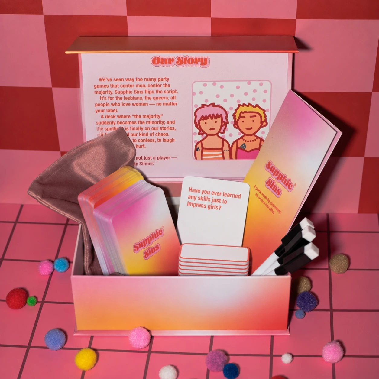Sapphic Sins open game box with cards, booklet, pouch and pens on a pink checkerboard set